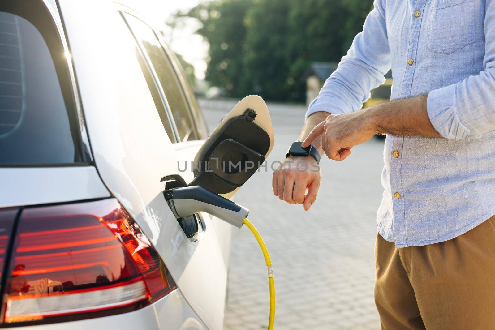 Male hand inserts power connector into EV car and charges batteries, uses smartwatch for activates start charging. Businessman plugging in charging cable to to electric vehicle.