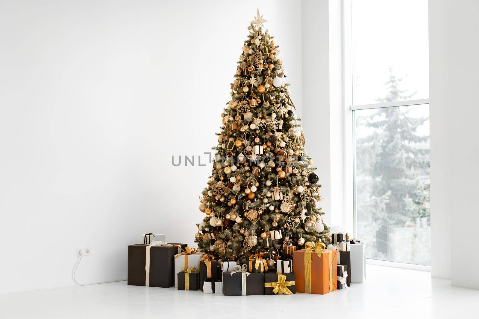 Christmas tree in white interior with stylish black and orange gift box. Minimalism. White wall background. Free space for text. Modern decorated Christmas fir Happy New Year. Holidays