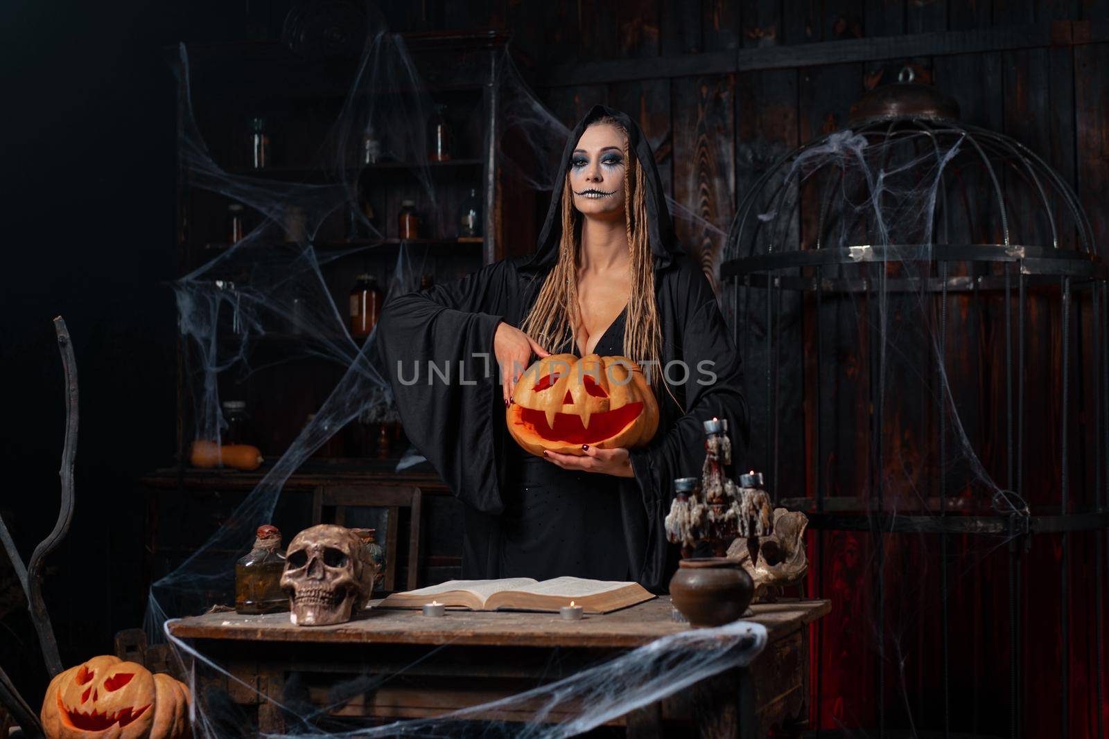 Halloween concept. Black witch holding Halloween pumpkin with carved smily face in hand standing dark room by andreonegin