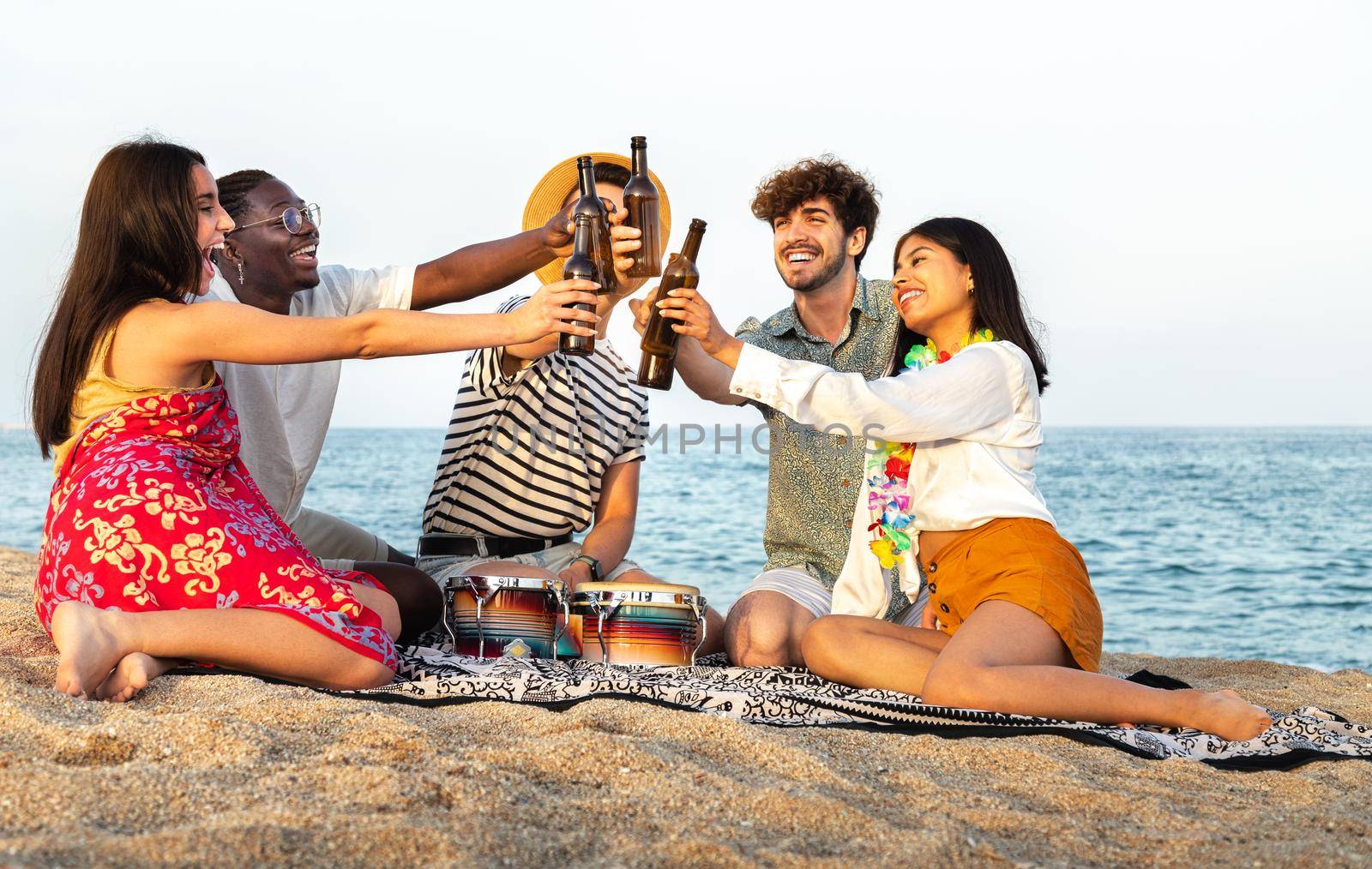 Happy, diverse group of friends toasting with beer together at the beach. Multiracial young people having fun outdoor. Vacation and friendship concept.
