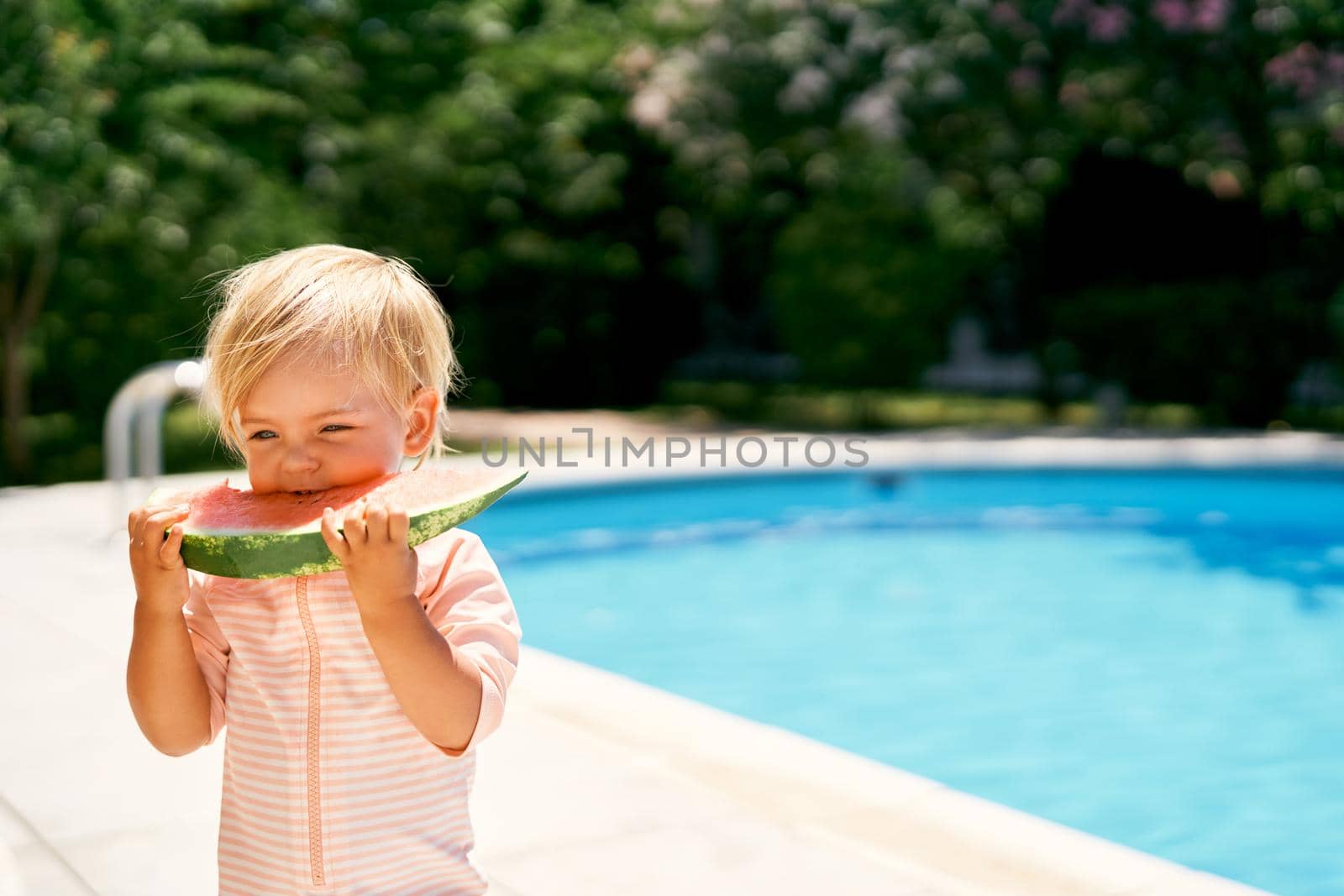 Little girl stands by the pool, eating a watermelon by Nadtochiy
