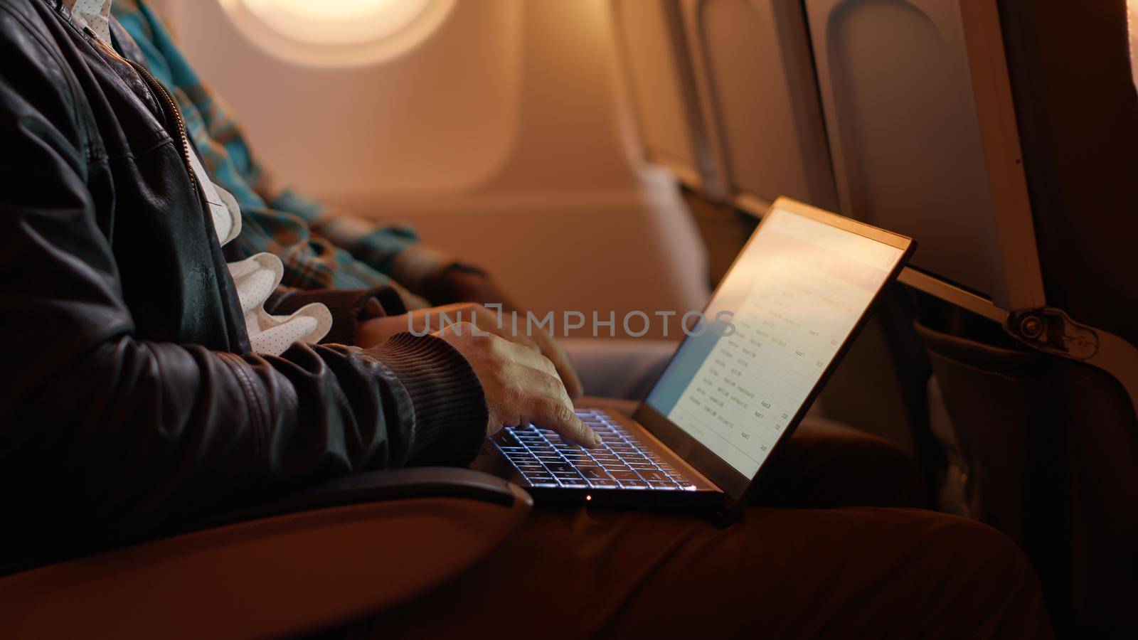 Tired employee travelling by airplane and working on laptop, using computer on flight during sunset. Work trip on plane, flying on vacation in economy class with people.