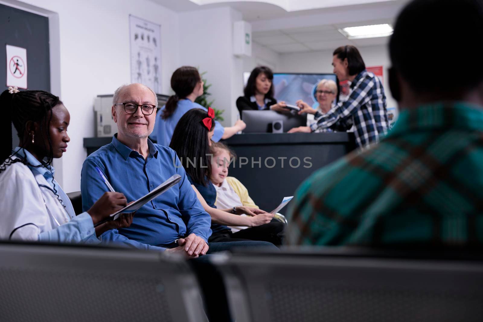 Portrait of older man with african american medical doctor looking at patient data while diverse people are talking at hospital reception. Senior citizen in busy hospital clinic attending appointment.