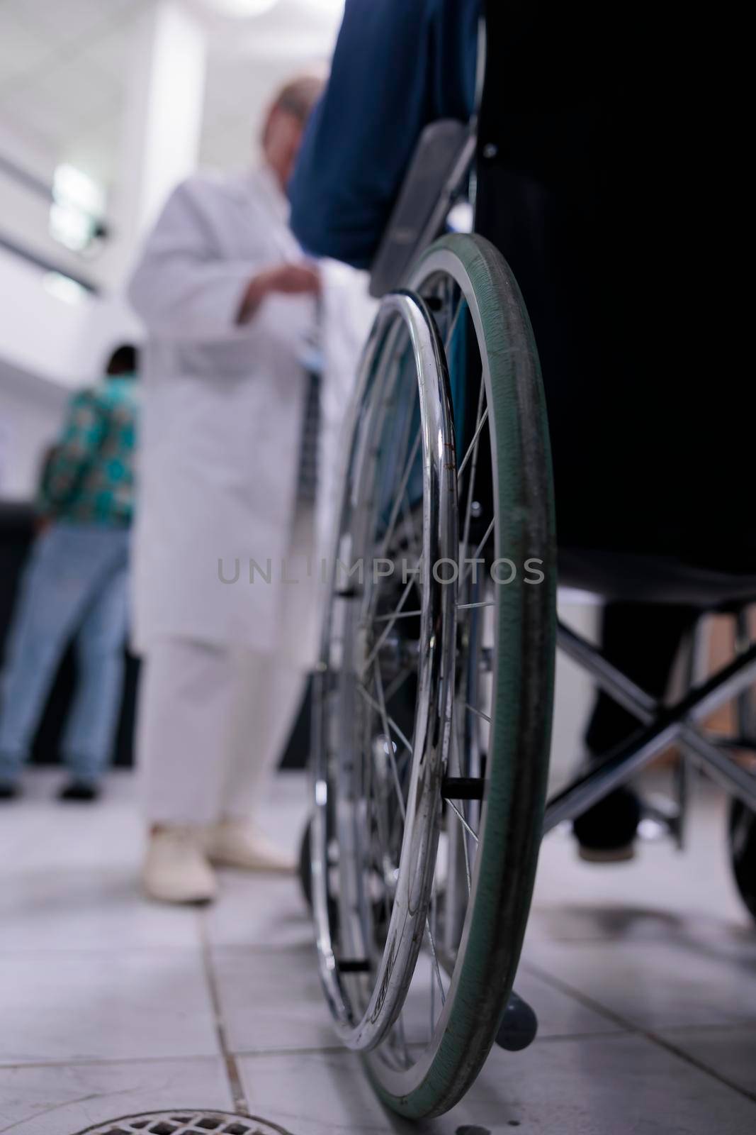 Closeup of senior man living with disability using wheelchair in private clinic reception talking with medical doctor about appointment. Selective focus on wheelchair wheel in busy hospital lobby.