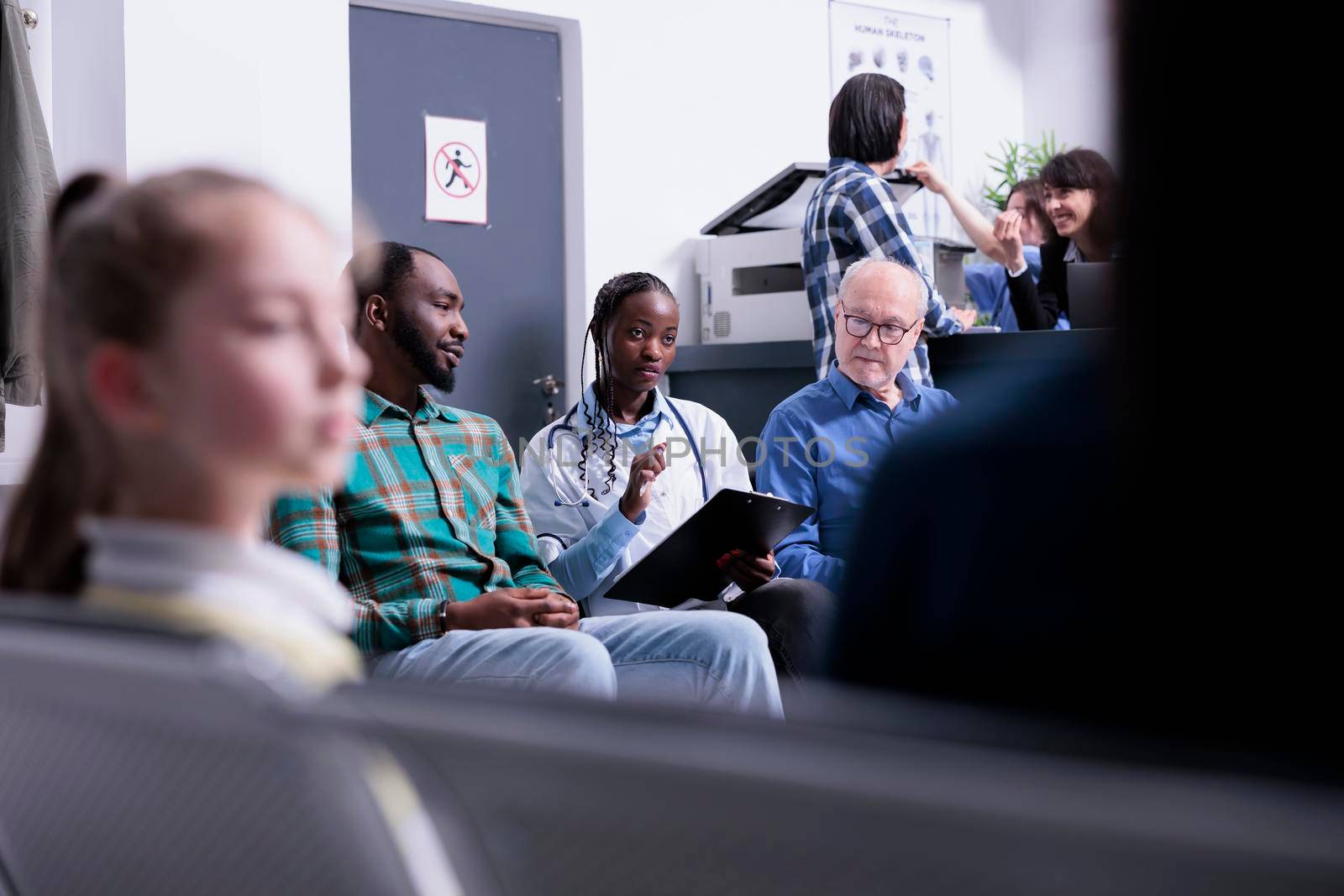 Selective focus on african american doctor with stethoscope completing form for patients in waiting room. Diverse people waiting in private clinic while hospital medic is asking medical questions.