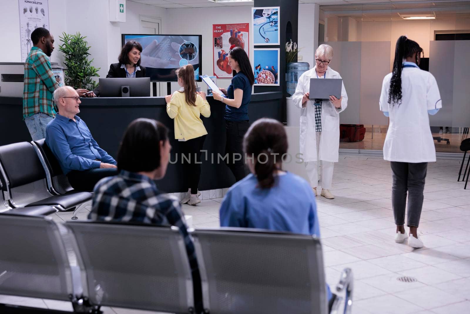 Senior doctor holding laptop computer in modern clinic reception while patients register for clinical consult by DCStudio