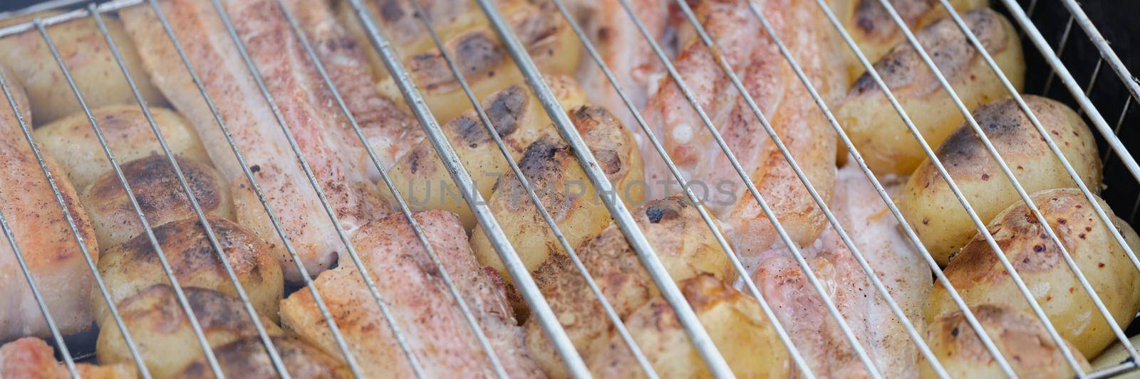 Close-up of fresh raw chicken legs and potatoes on grill gridiron in nature. Picnic on fresh air, frying meat and vegetable. Street food, bbq or picnic concept