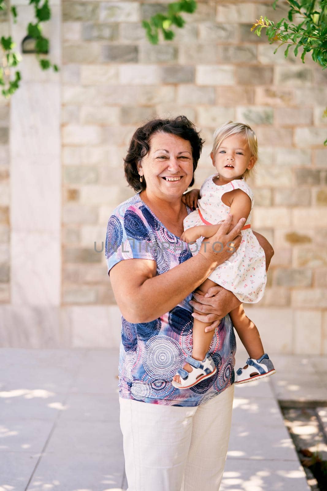 Grandmother stands holding a little girl in her arms near a stone wall by Nadtochiy