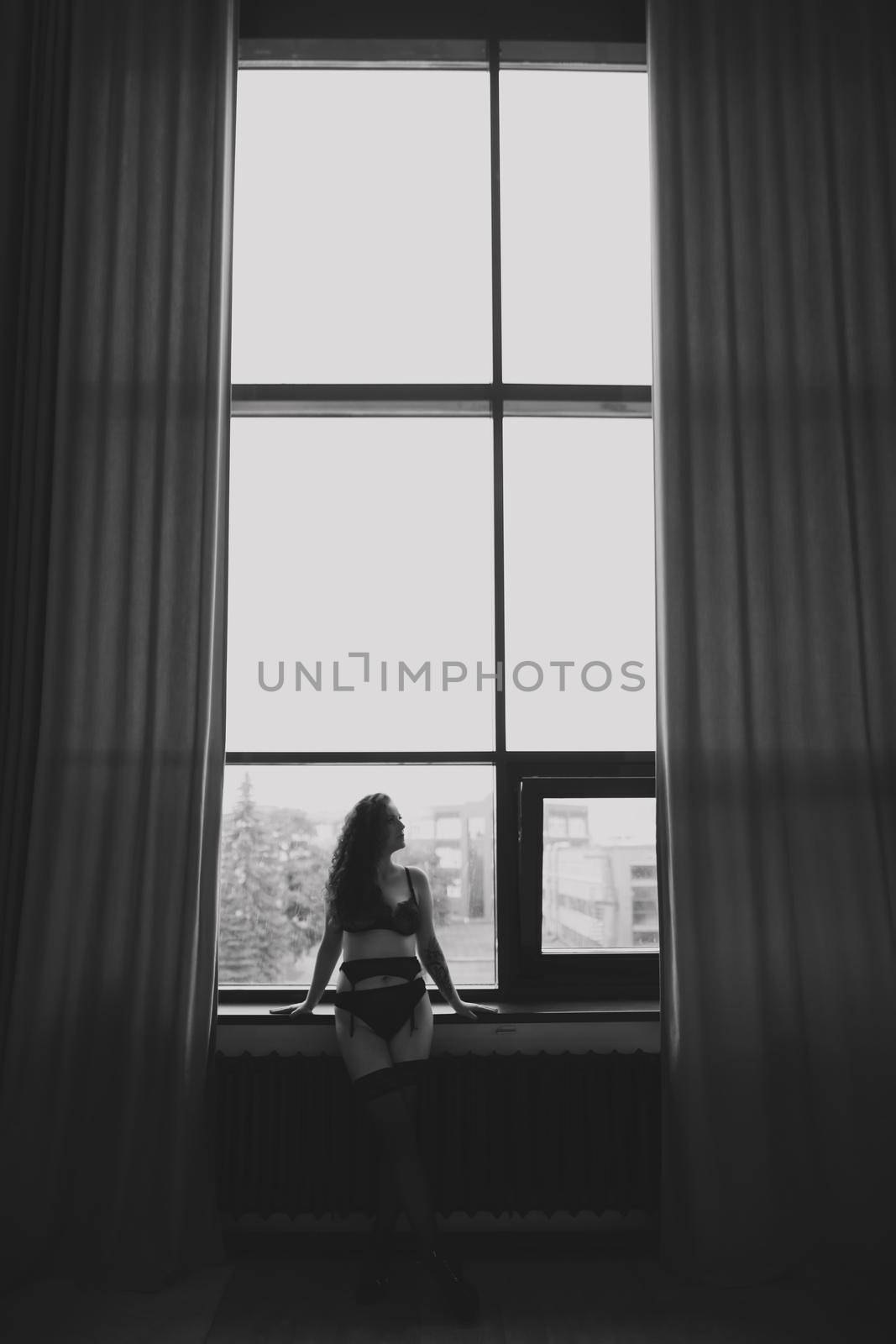 Gorgeous woman wearing black lingerie indoors. Portrait of a beautiful dreamy girl in a black underwear by the window. Black and white photo