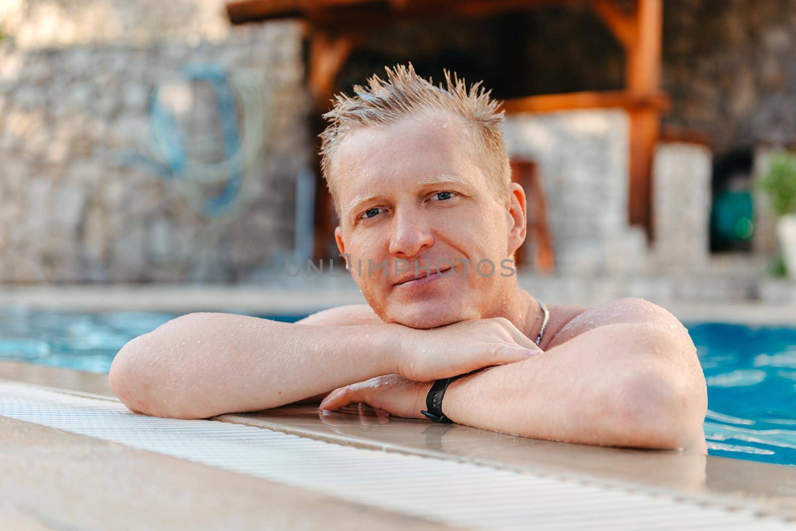 Photo of handsome smiling man in swimming pool in summer scenery. Attractive, cheerful, stunning, perfect, harsh, virile man in yellow shorts, having cocktail with small umbrella in hand and holding arm in pocket, isolated on blue background