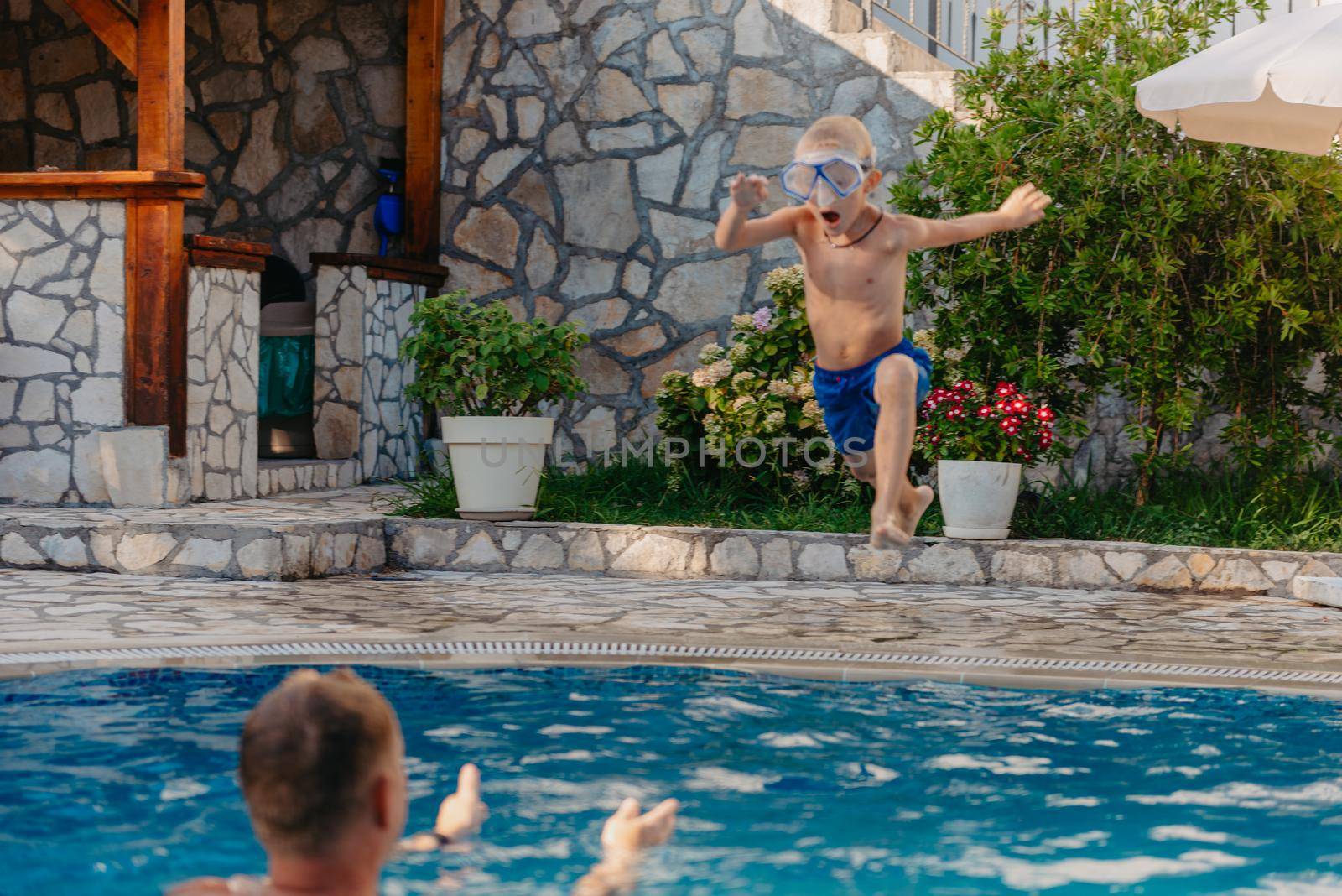 Excited boy in googles jumping in water from shoulders of his father standing in swimming pool