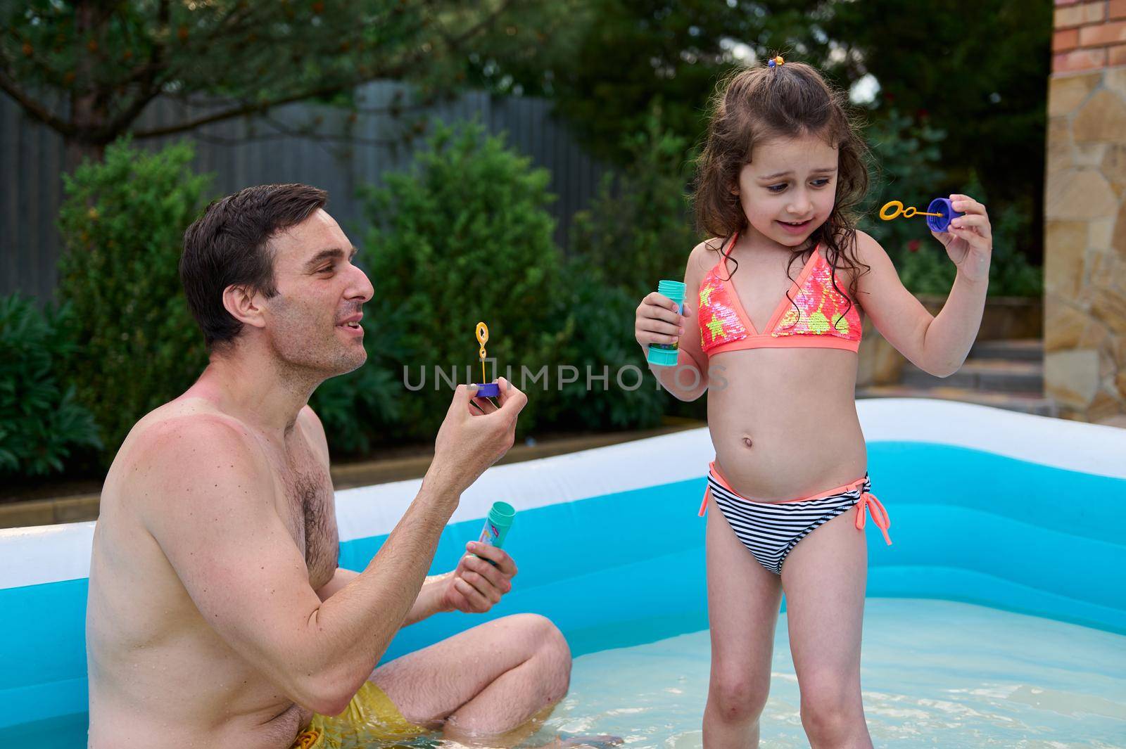 Adorable child, baby girl in bathing suit blows soaps bubbles in the inflatable swimming pool, next to her loving farher. Happy childhood. Family weekend together, love, togetherness. Father's Day