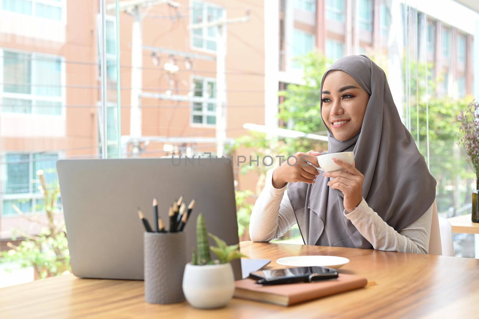 Thoughtful Muslim businesswoman drinking coffee and looking through glass office window, enjoying city view.