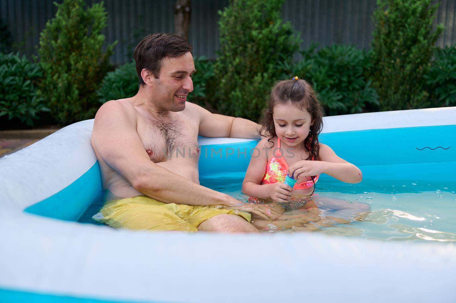 Pleasant middle-aged Caucasian man, loving father with his cute daughter, a charming little girl blowing soap bubbles while having fun in the inflatable water pool at home garden on a sunny summer day