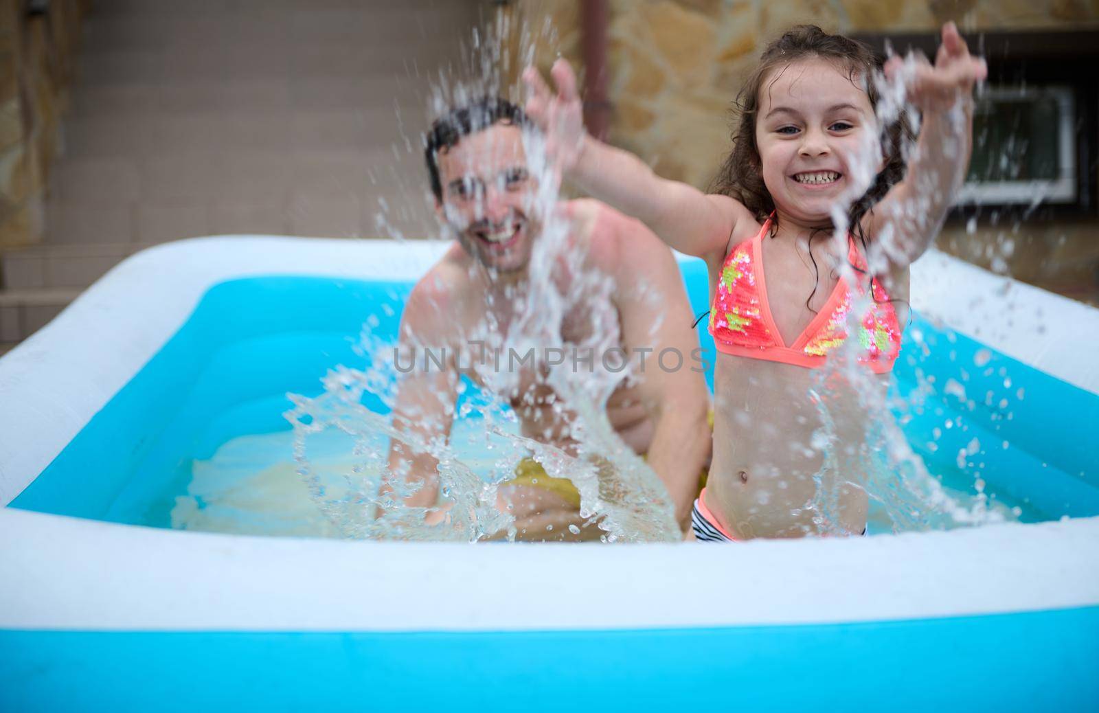 Cheerful little girl and her loving dad splashing water, looking at camera while having fun together in the inflatable swimming pool outdoors. Father and daughter. Happy and carefree childhood. Family