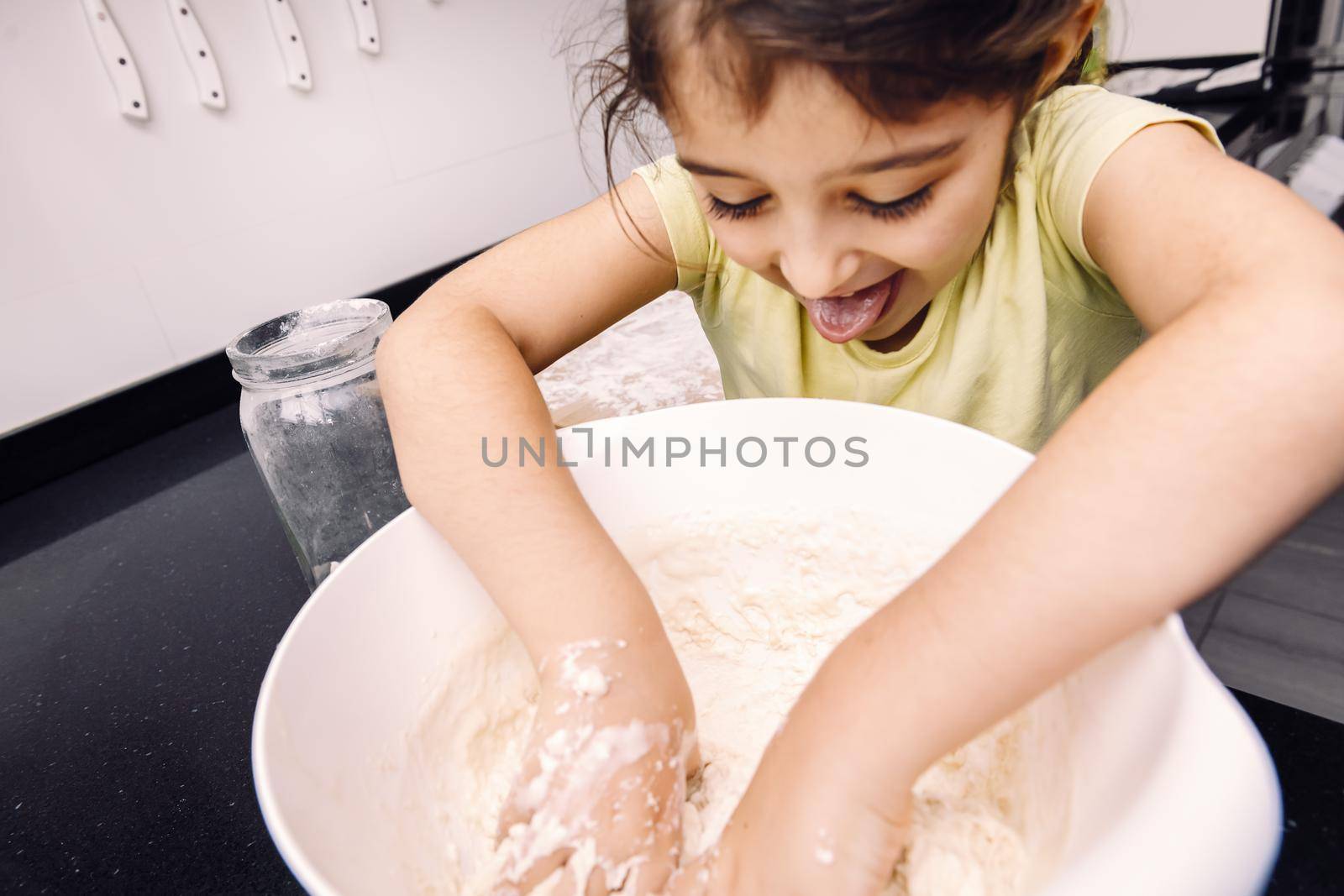 funny little girl with her tongue out mixing flour in a bowl to make homemade bread dough, healthy food concept at home