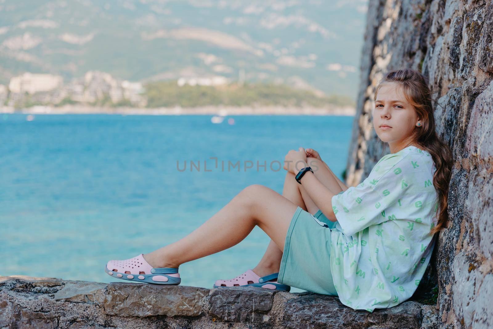 Beautiful girl sitting on a stone wall, in background is the blue sea, Budva, Montenegro. by Andrii_Ko