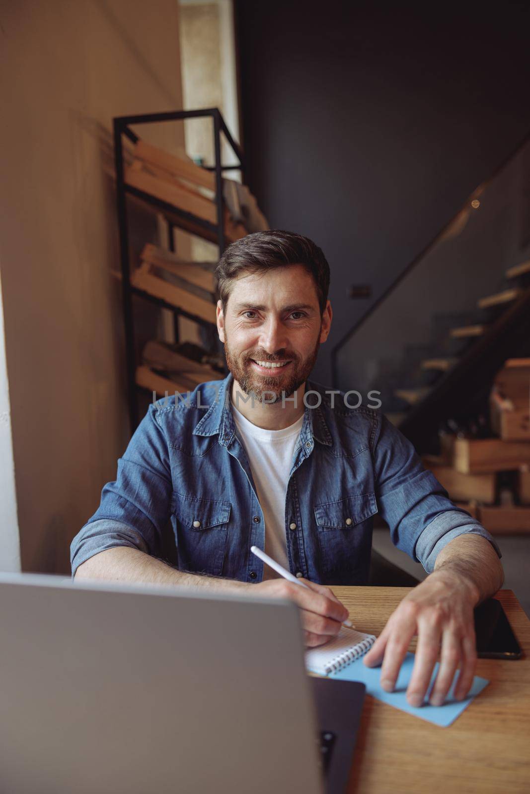 Portrait of handsome man sitting in cafe at laptop with pen and notebook and smiling. Vertical shot.
