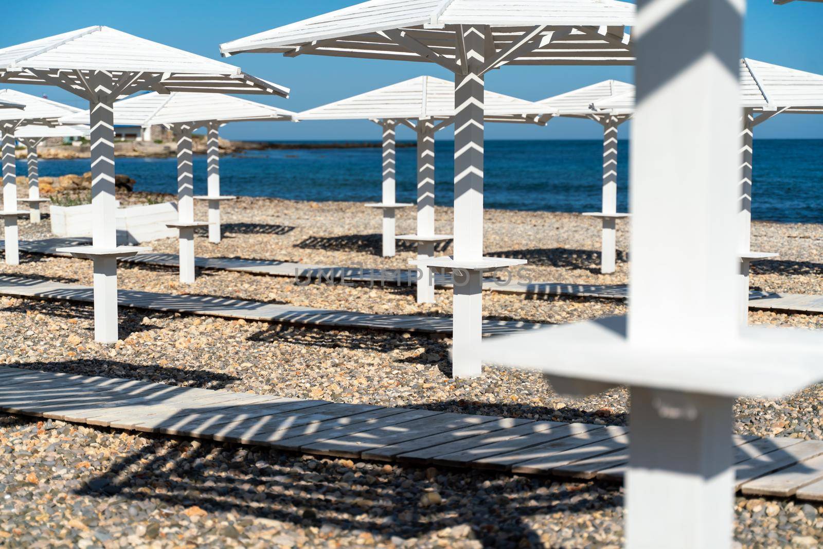 Rows of wooden umbrellas from the sun on the seashore in the morning. Wooden paths on the sand between umbrellas. Beach holiday at the resort. by Matiunina