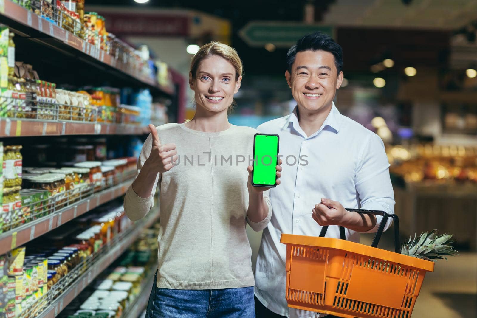 Portrait of a happy Asian couple of consumers, shoppers in a supermarket or grocery store, looking at the camera, smiling. are holding a phone and showing a green chromakey screen