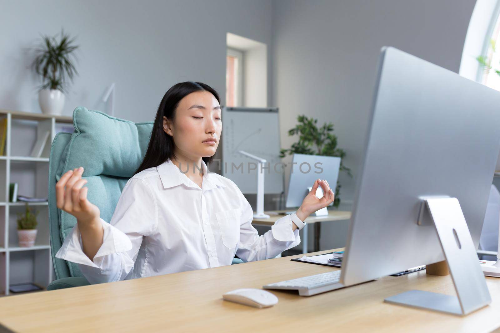 Young asian businesswoman sitting in office with eyes closed, holding hands in lotus yoga pose by voronaman