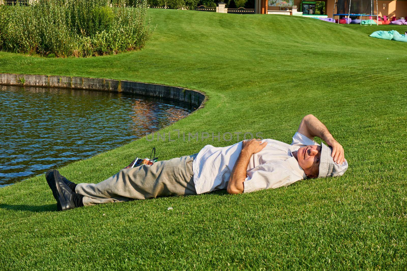 An elderly man is resting on a mowed lawn in a large field near a pond by jovani68