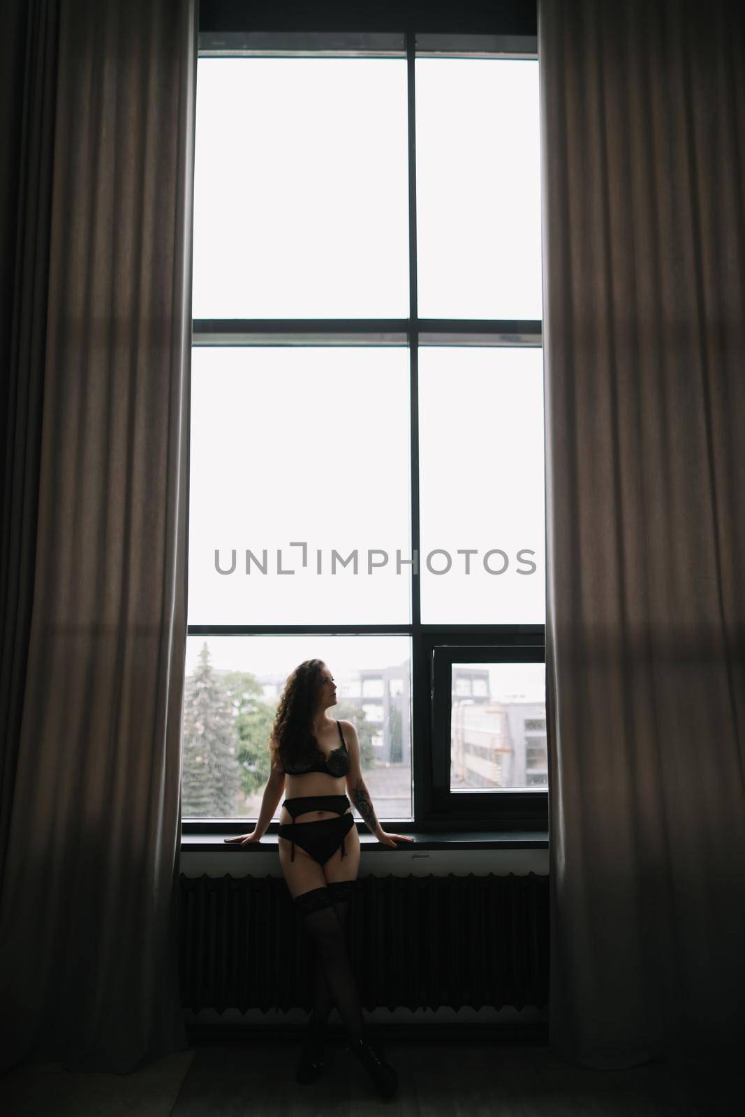 Gorgeous woman wearing black lingerie indoors. Portrait of a beautiful dreamy girl in a black underwear by the window. Black and white photo