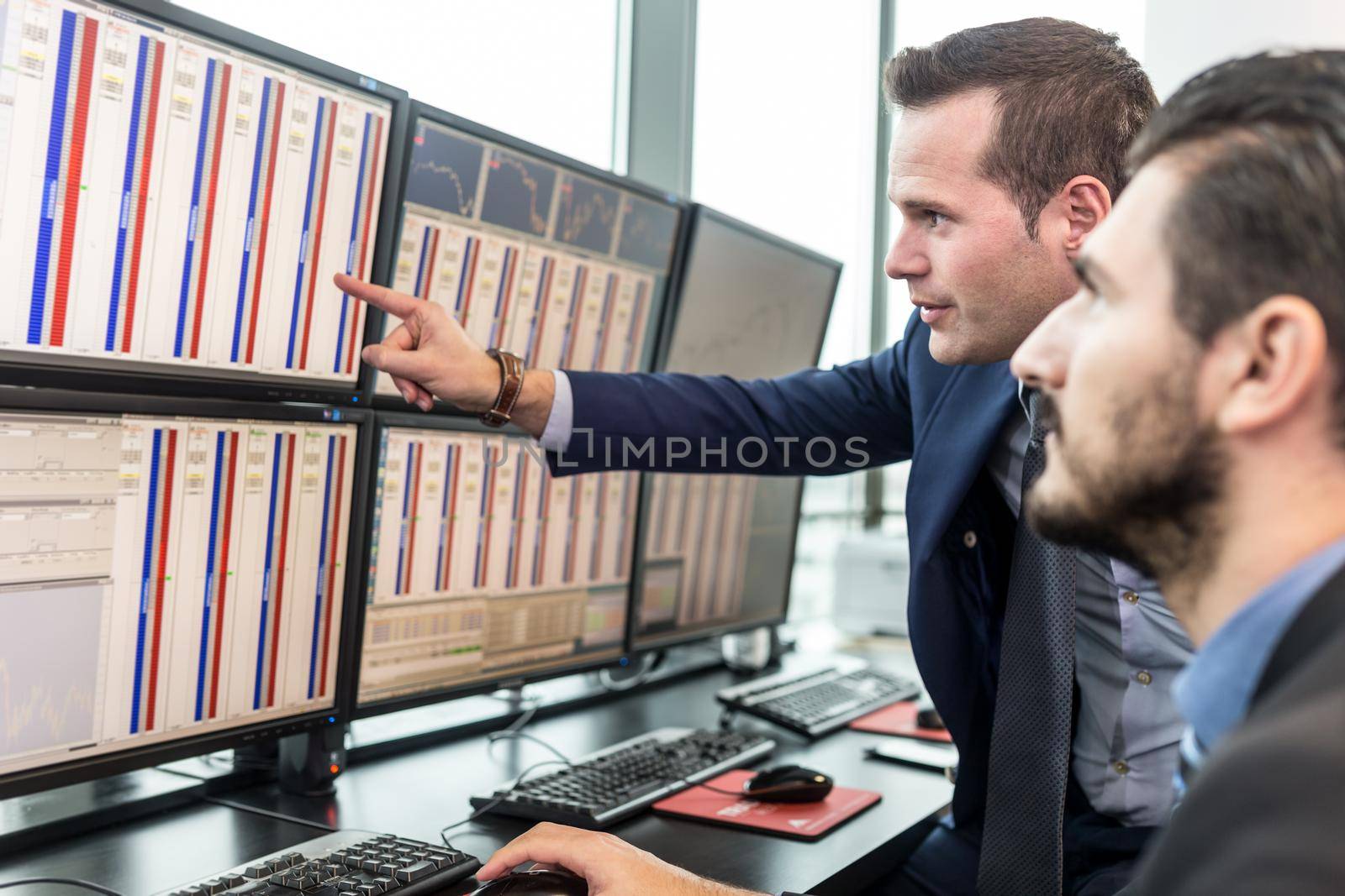 Businessmen trading stocks. Stock traders looking at charts, indexes and numbers on multiple computer screens. Colleagues in discussion in traders office. Broker pointing on market data on monitor.