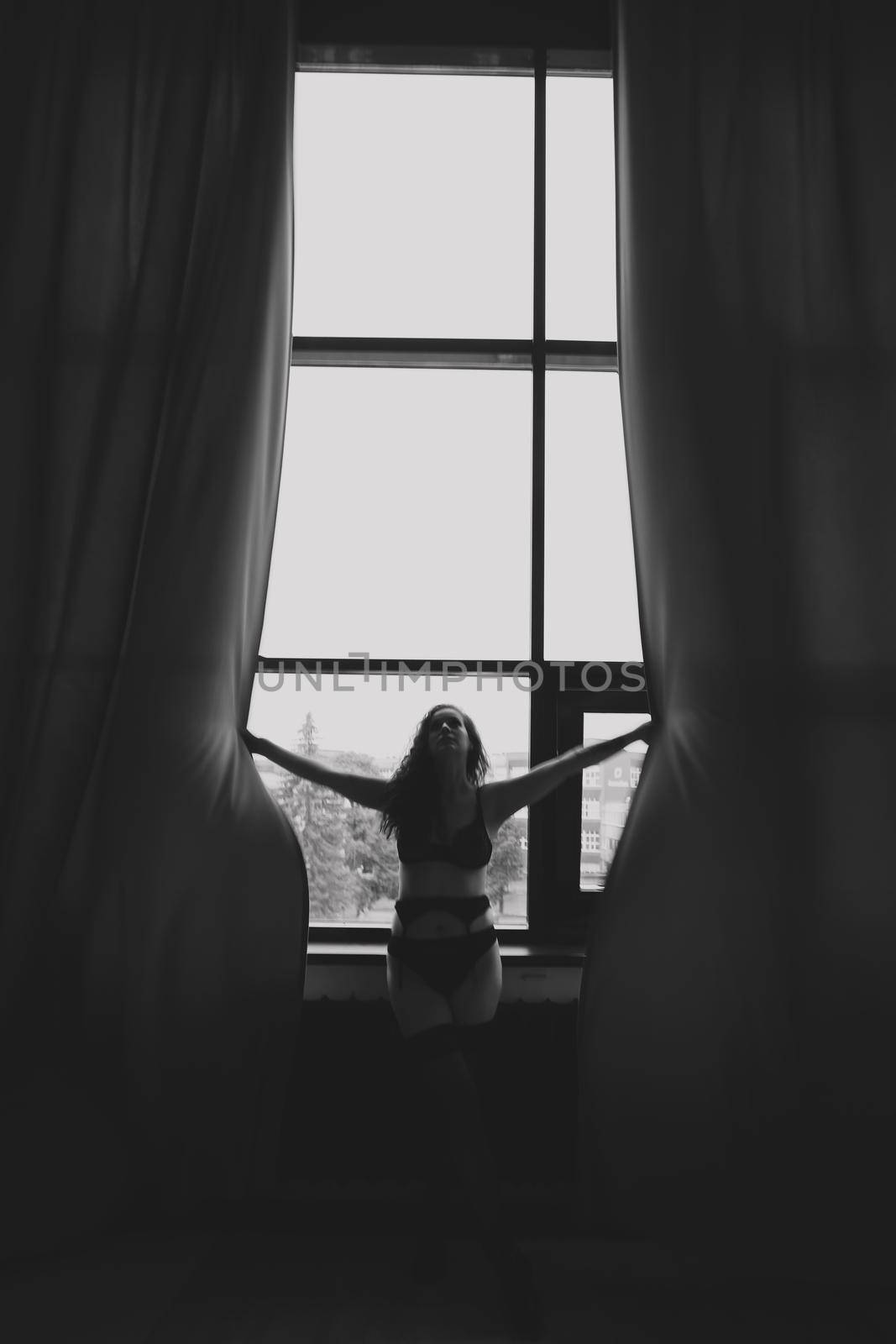Gorgeous woman wearing black lingerie indoors. Portrait of a beautiful dreamy girl in a black underwear by the window. Black and white photo