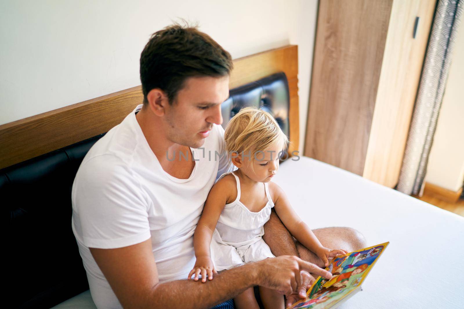 Dad reads a book with colorful pictures to a little girl on the bed. High quality photo