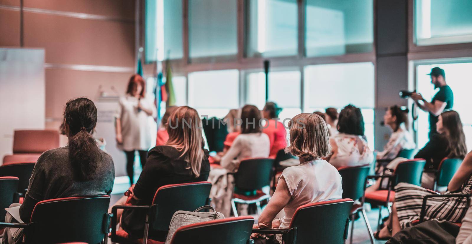 Business and entrepreneurship symposium. Female speaker giving a talk at business meeting. Audience in conference hall. Rear view of unrecognized participant in audience.