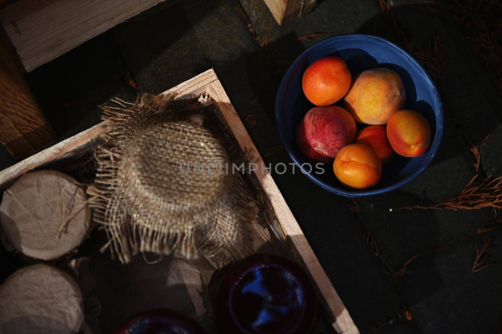 Focus on a blue ceramic bowl of ripe, juicy and ready-to-eat apricots from organic garden, nearby a wooden crate with homemade fruit jam jars. Top view. Copy ad space. Healthy food and canning concept