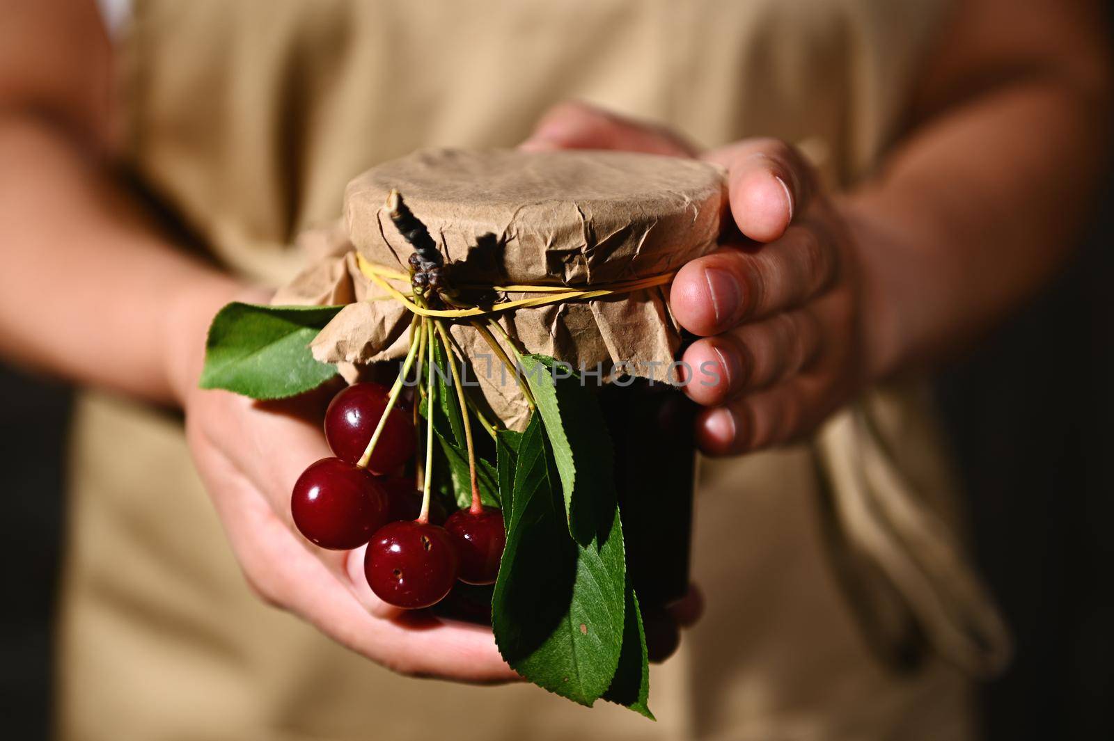 Details: Hands of a woman confectioner in a beige chef apron, handing a jar of homemade jam of ripe fresh cherries. Canning, preserved sweet food.