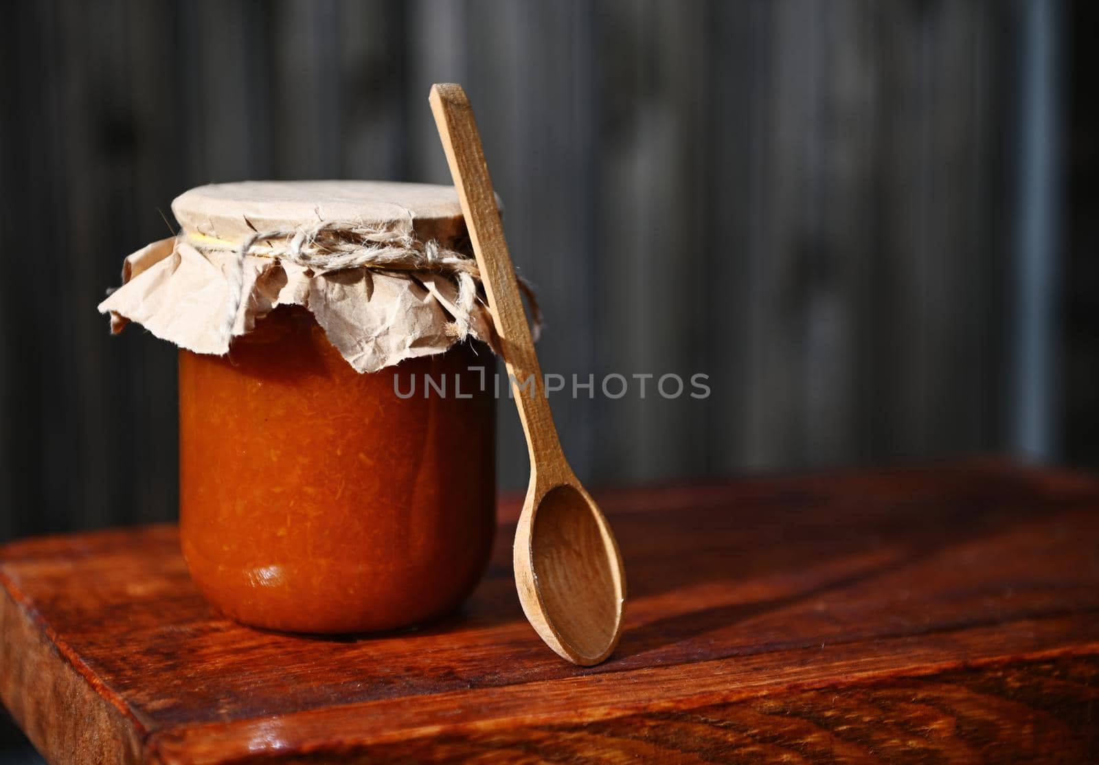Still life of a glass jar of apricot jam, with craft paper on the lid tied with rope and a wooden tea spoon on a rustic wood surface with copy ad space for text. Canning and preserved food concept