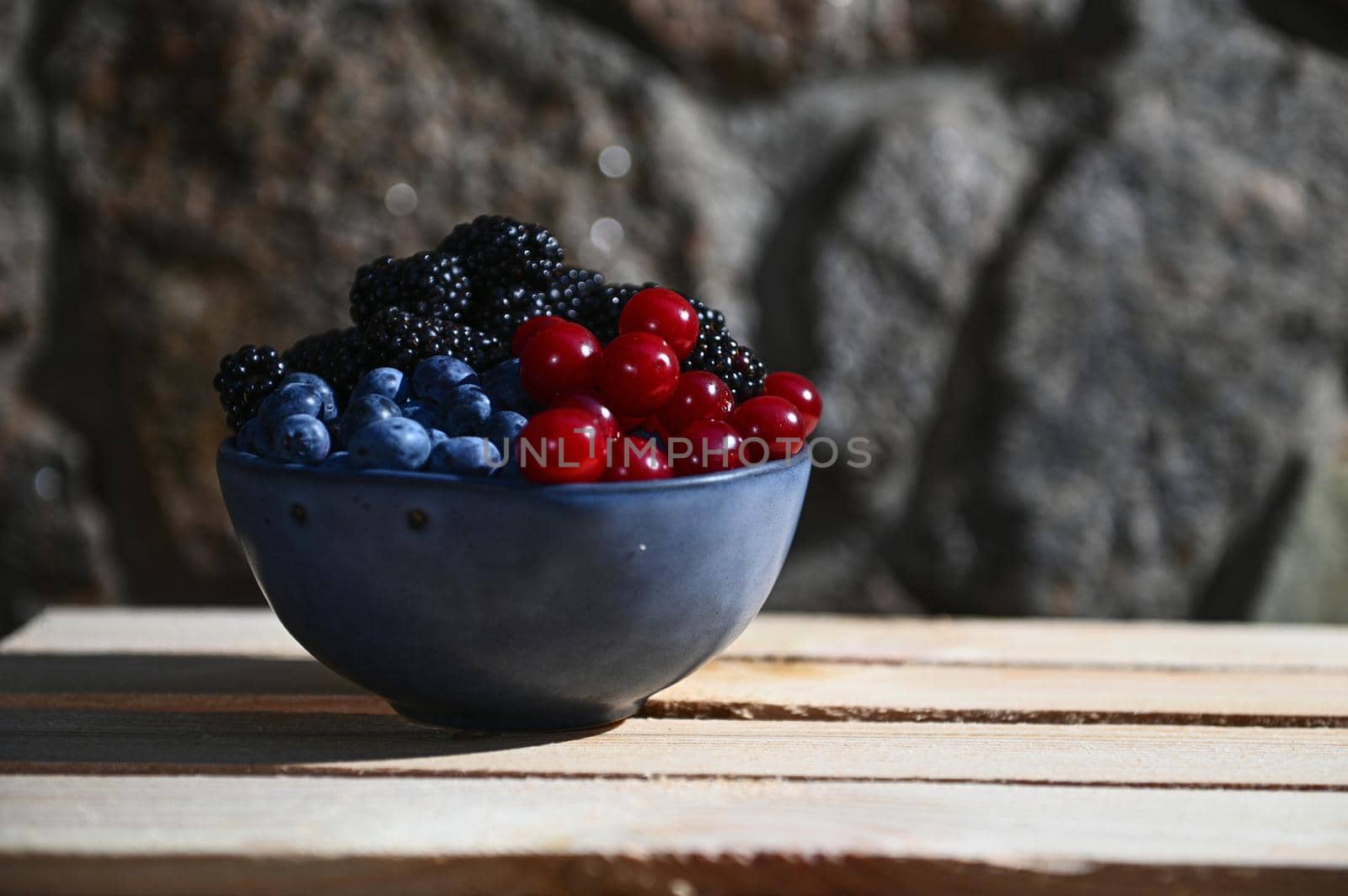 Close-up of fresh juicy and ripe seasonal wild berries from an organic farm- blueberries, blackberries and cherries in a blue ceramic bowl on a wooden crate against a gray stone backdrop. Healthy food
