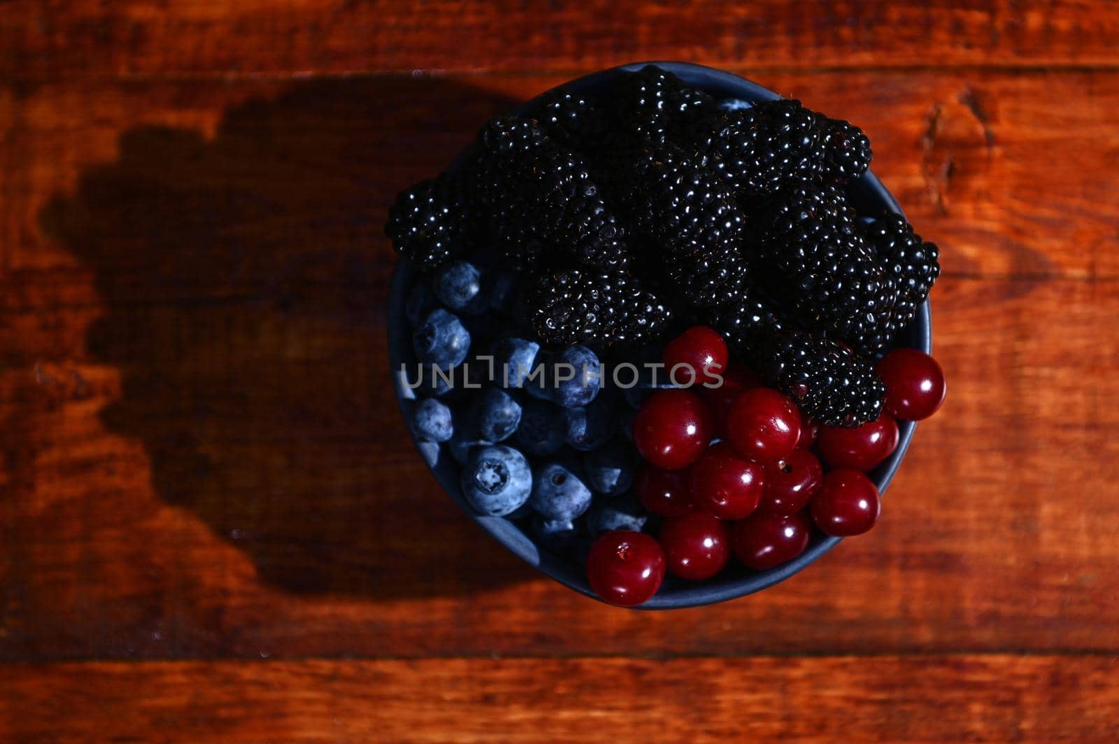 Flat lay. Blue ceramic bowl of fresh, ripe and ready-to-eat organic wild berries- blueberries, blackberries and cherries from and organic farm, on a wooden background. Copy ad space for text. Banner