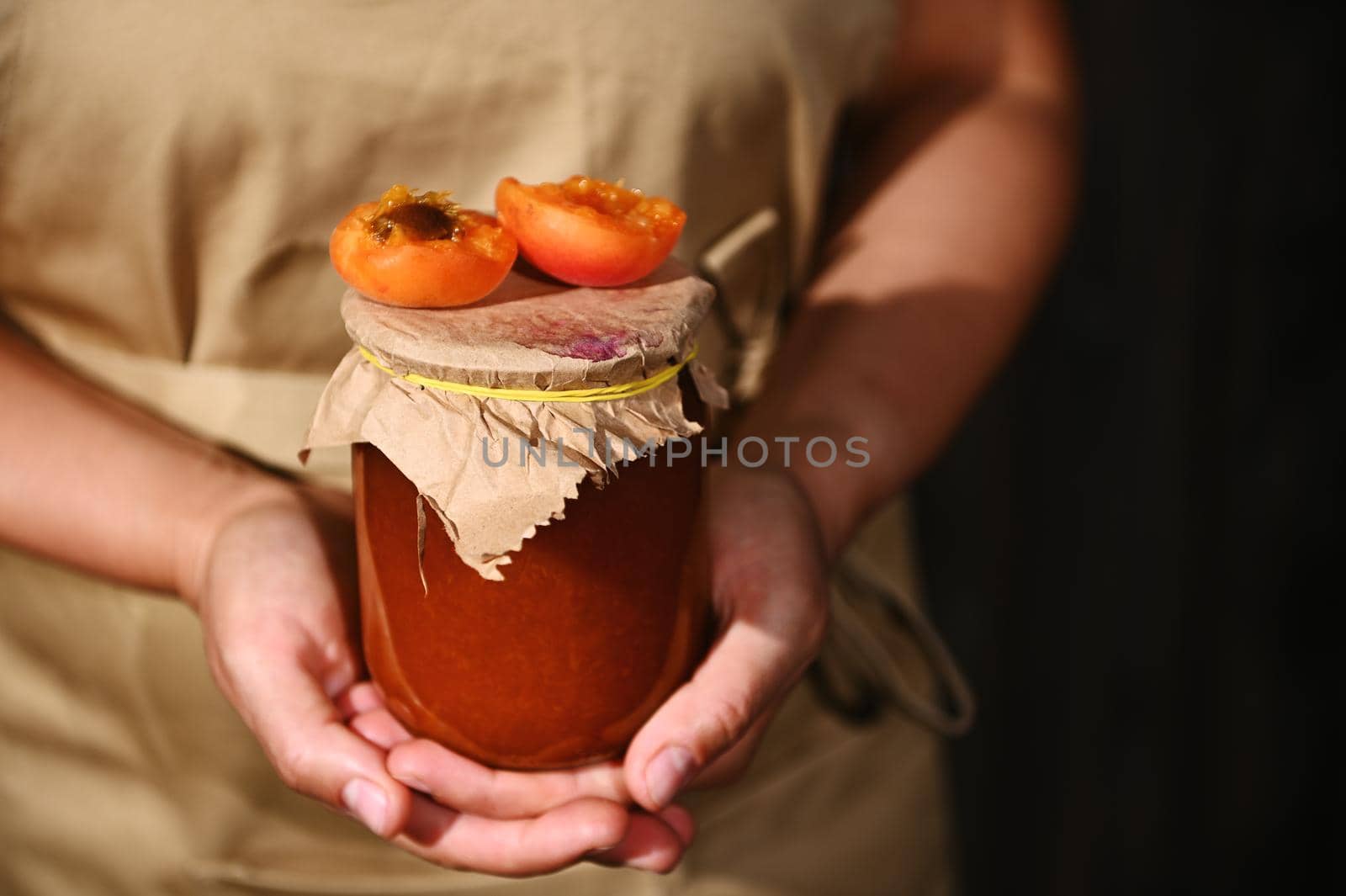 Details: Hands of a woman confectioner in a beige chef apron, holding a jar of homemade jam with juicy ripe red summer sunny apricot broken in halves on the craft lid. Canning, preserved sweet food.