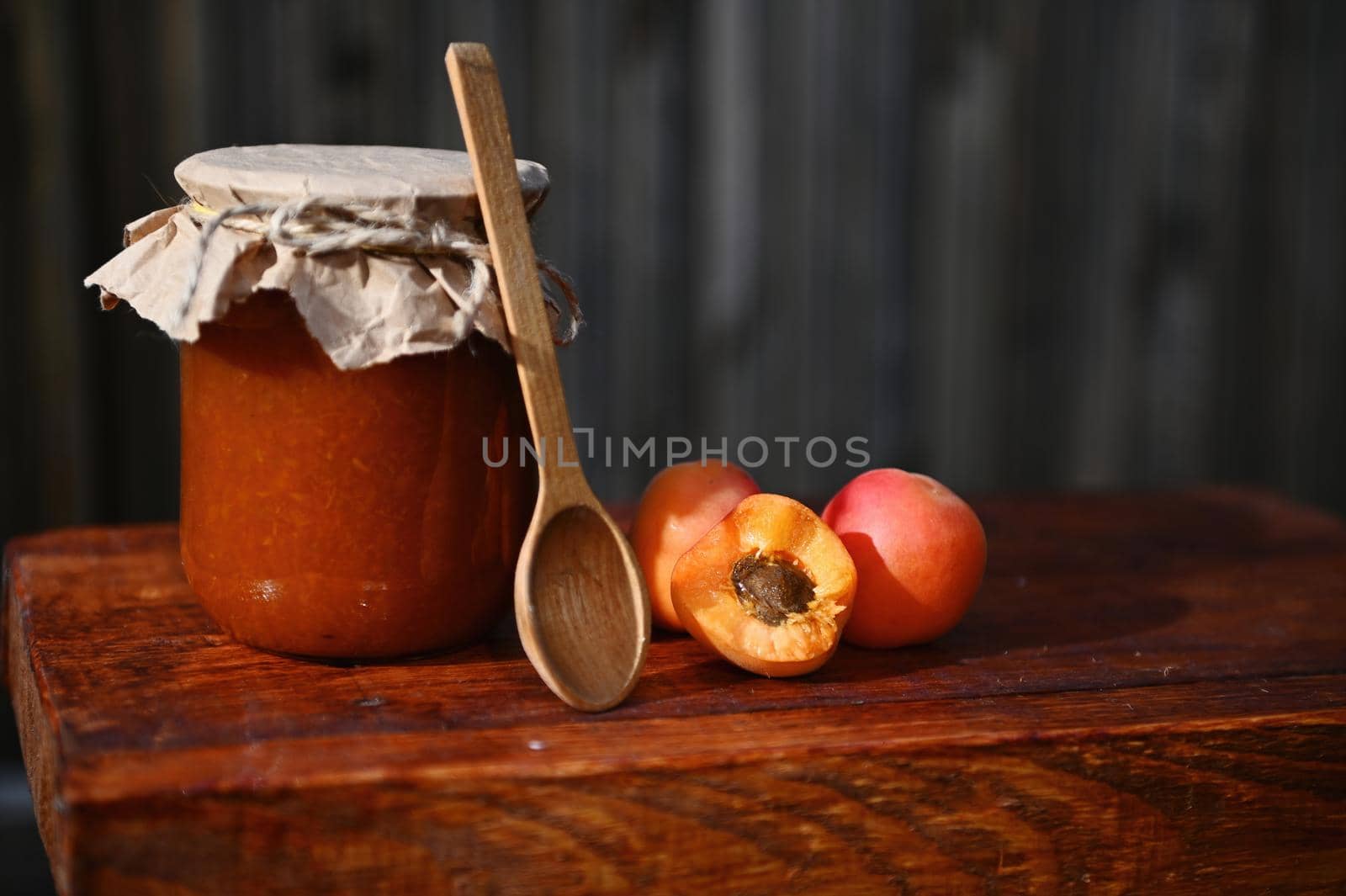 Still life. A glass jar of homemade jam, with craft paper on the lid tied with rope, halves of ripe ready-to-eat red sunny apricots and wooden spoon on rustic wood surface. Canning, sweet preserves.