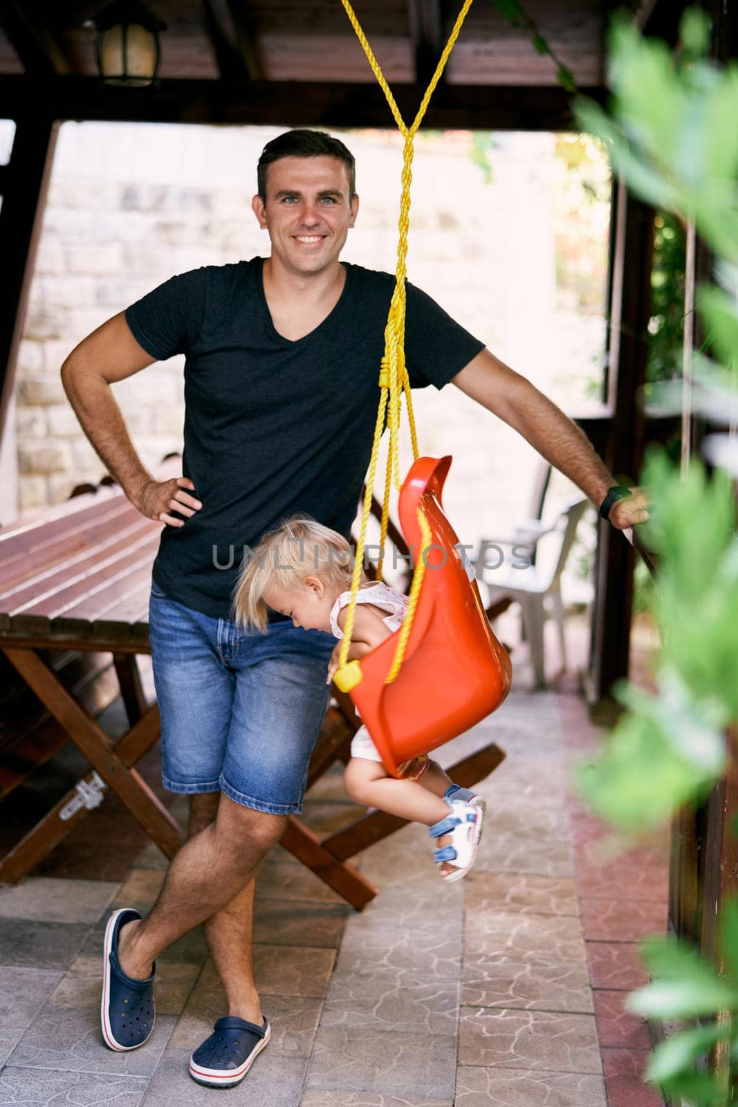 Dad stands next to a little girl on a swing in a wooden gazebo by Nadtochiy