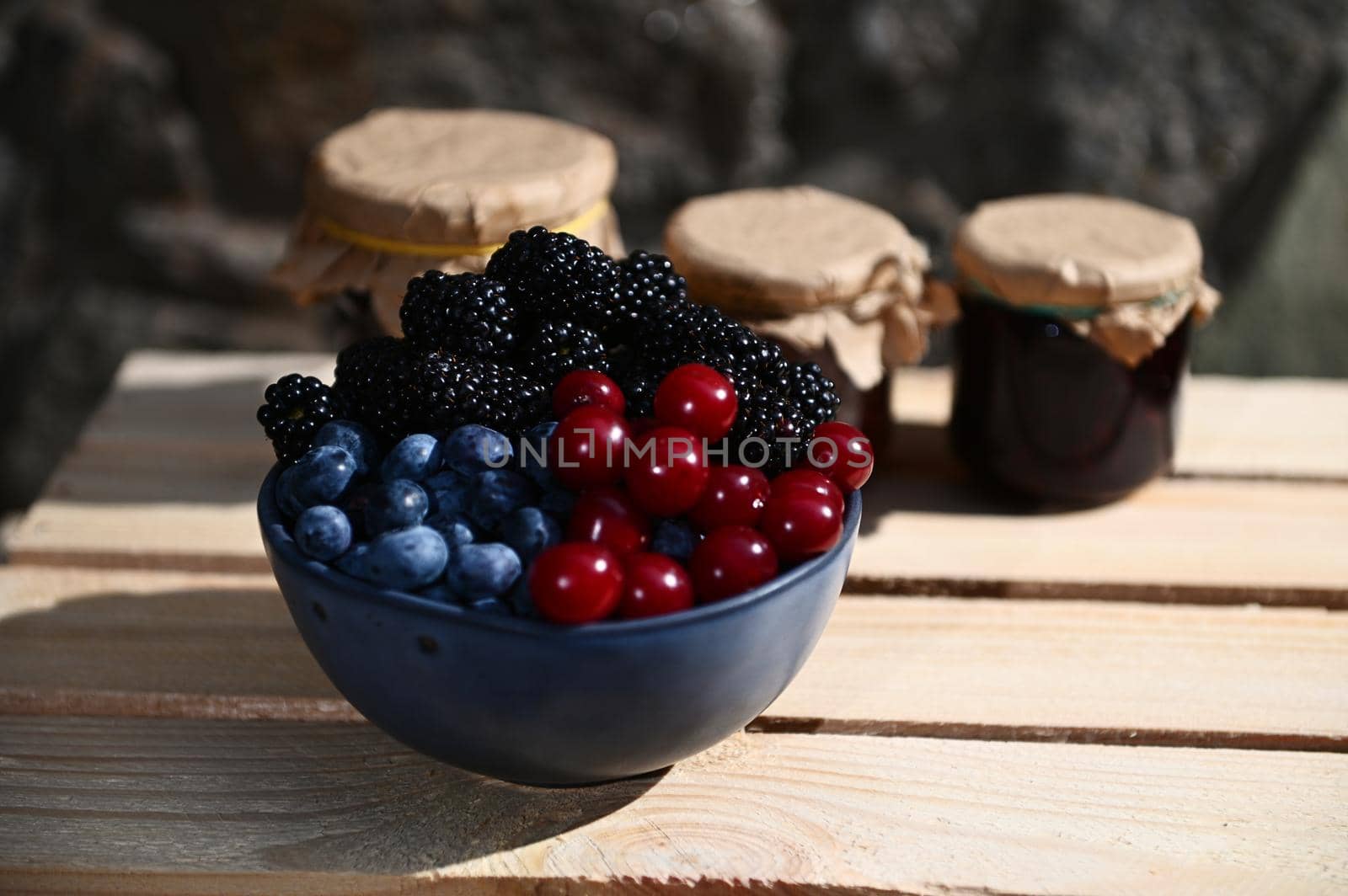 Close-up of a blue ceramic bowl with ripe cherry berries, blueberries and blackberries in a wooden box against the background of blurry assorted fruit jams in jars. Organic farm and canning concept