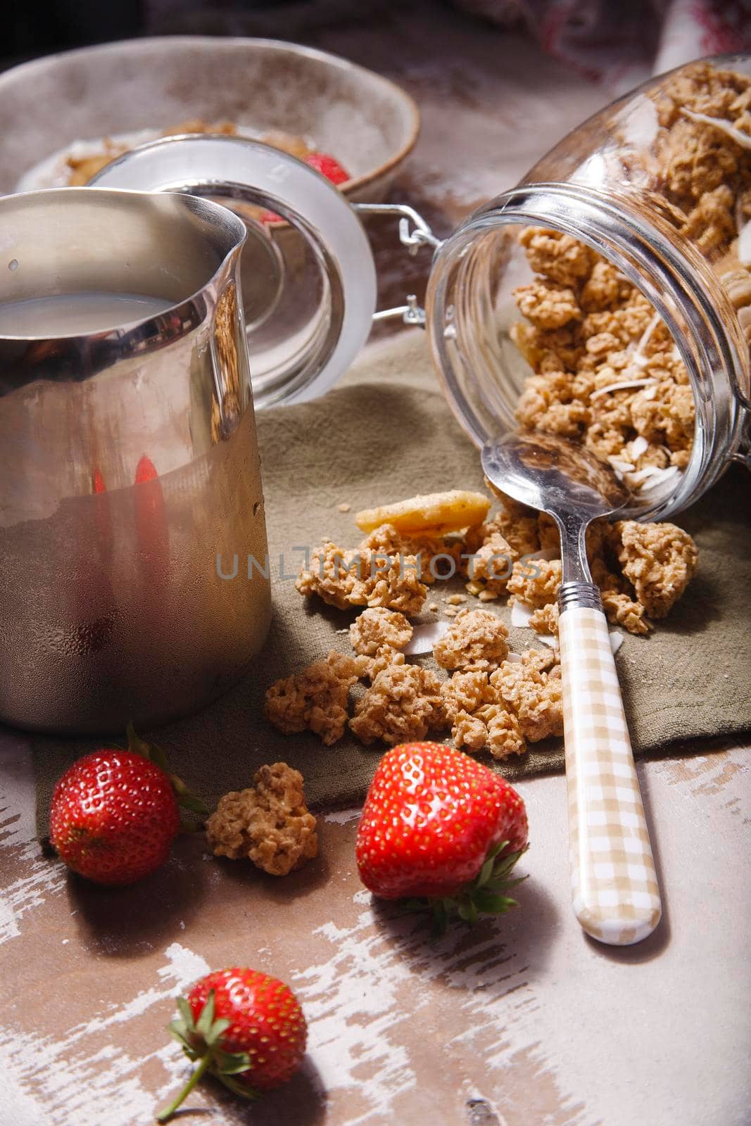 Homemade scattered granola in glass jar with fresh strawberry and milk on wooden table, selective focus