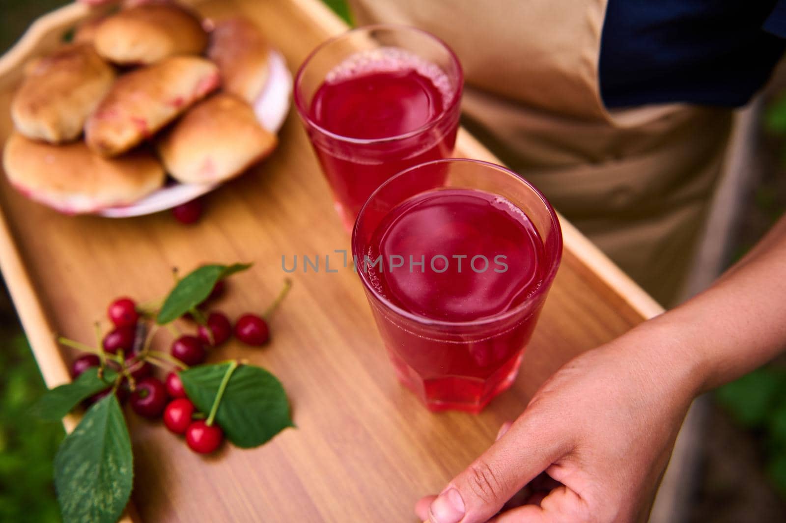 Focus on a cherry berry compote in drinking glasses near a dish with homemade freshly baked cherry pies and ripe juicy cherries on the serving tray in the hands of an unrecognizable woman, housewife