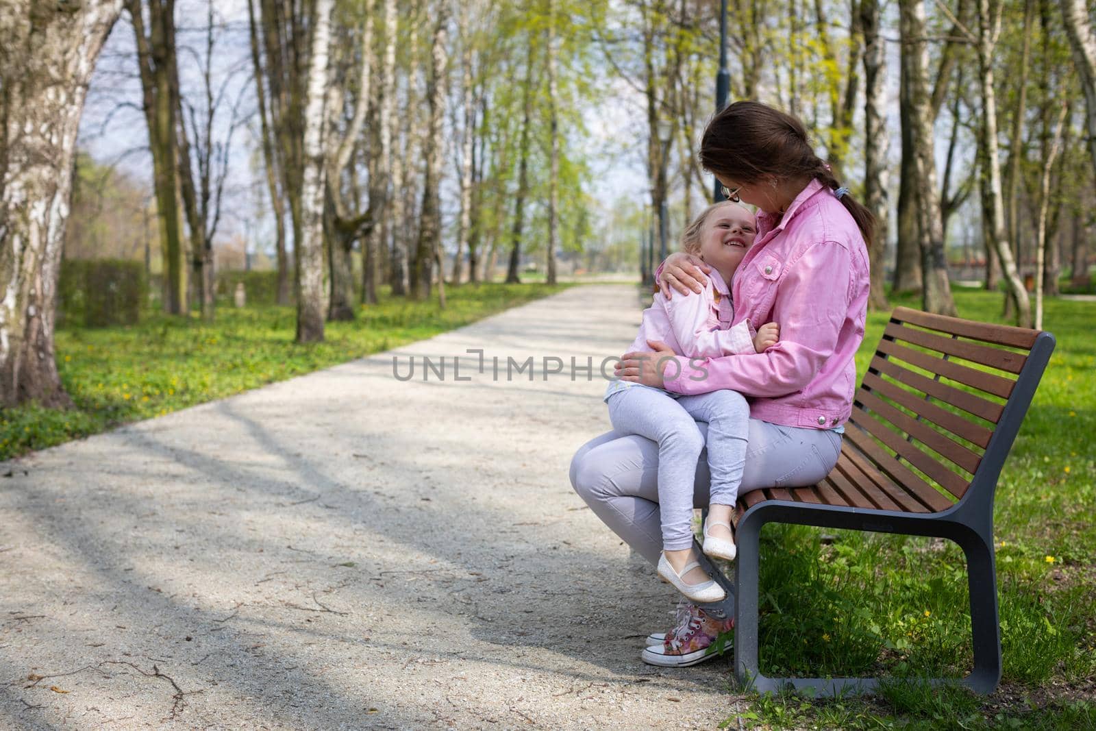 On a spring morning in the park, a mother and her daughter sit on a bench. The four-year-old girl is smiling. The mother holds the child in her lap on a sunny day.