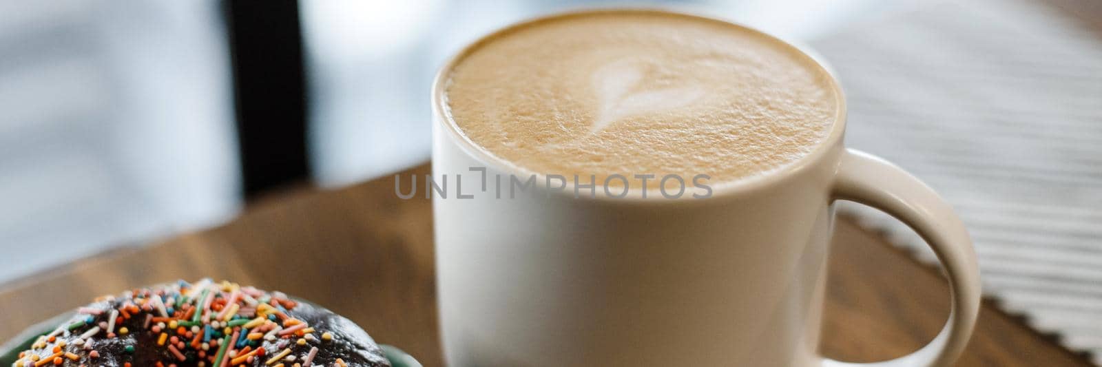 Coffee with a drawn heart and milk on a wooden table in a coffee shop. Pink donut with scattering on the table next to the coffee. Web banner.