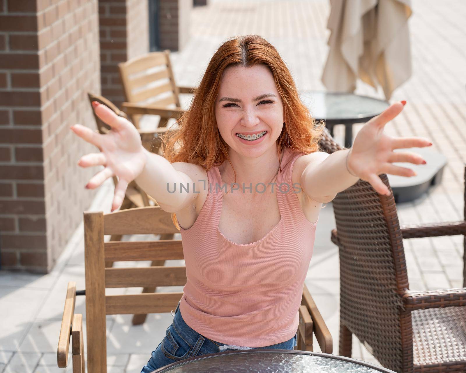 Young red-haired woman sits in a street cafe and stretches her arms to hug