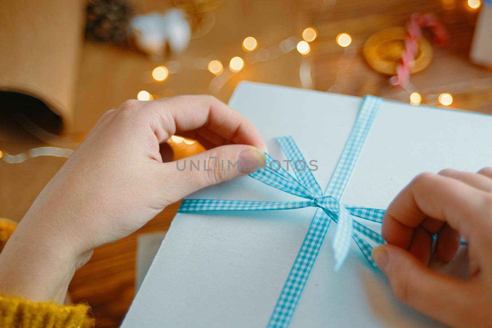 Women's hands tie blue ribbon bow on white gift box. Surprise for Birthday or Christmas. New Year's mood. Garland lights and decorations in background.