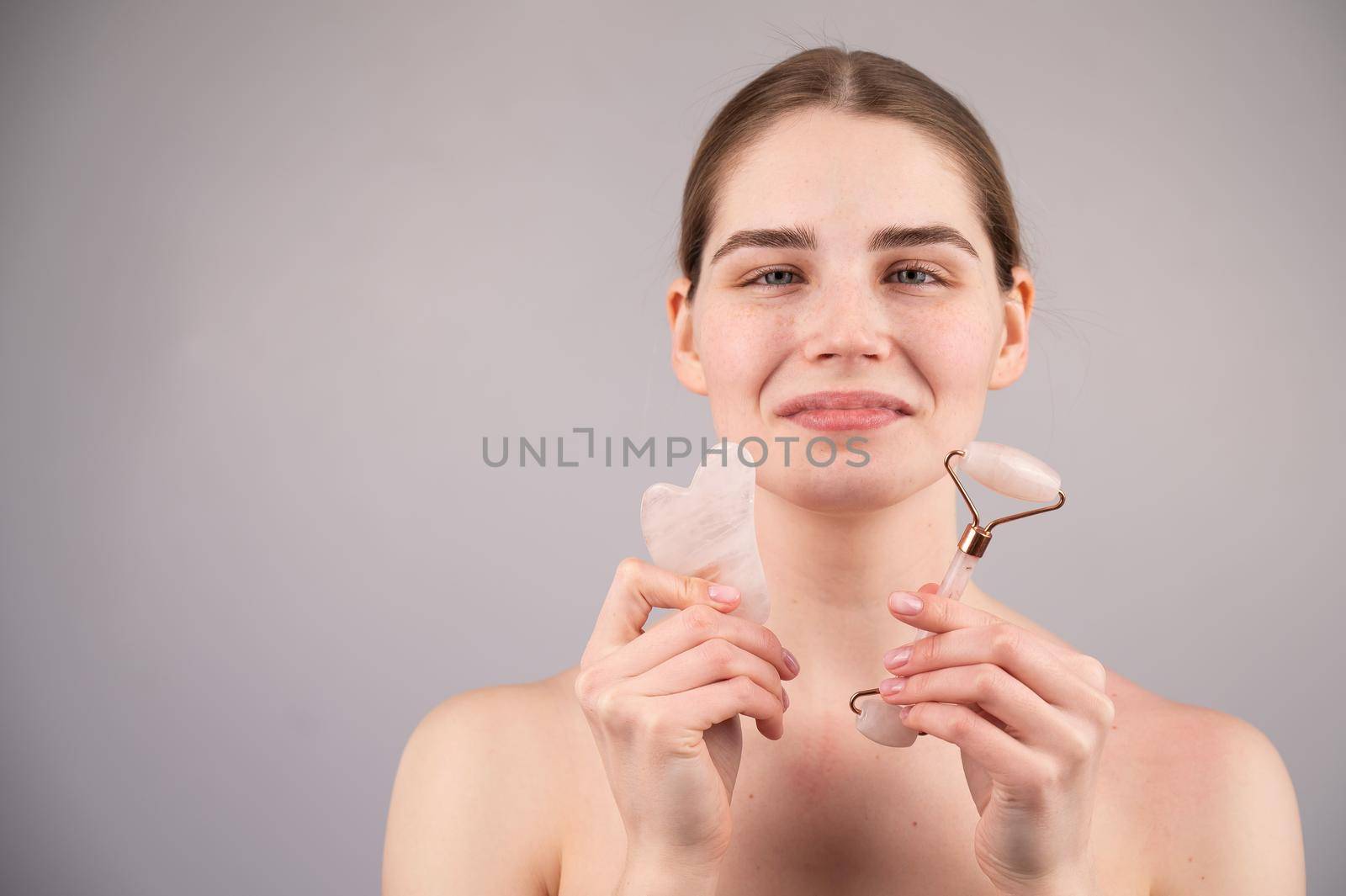 Caucasian woman holding pink roller massager and gouache scraper on white background. Copy space