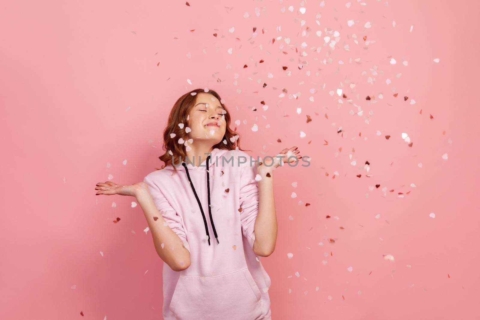 Extremely happy young brunette female enjoying falling heart shaped confetti and smiling with closed eyes, celebrating valentines day, holiday mood. Indoor studio shot, isolated on pink background