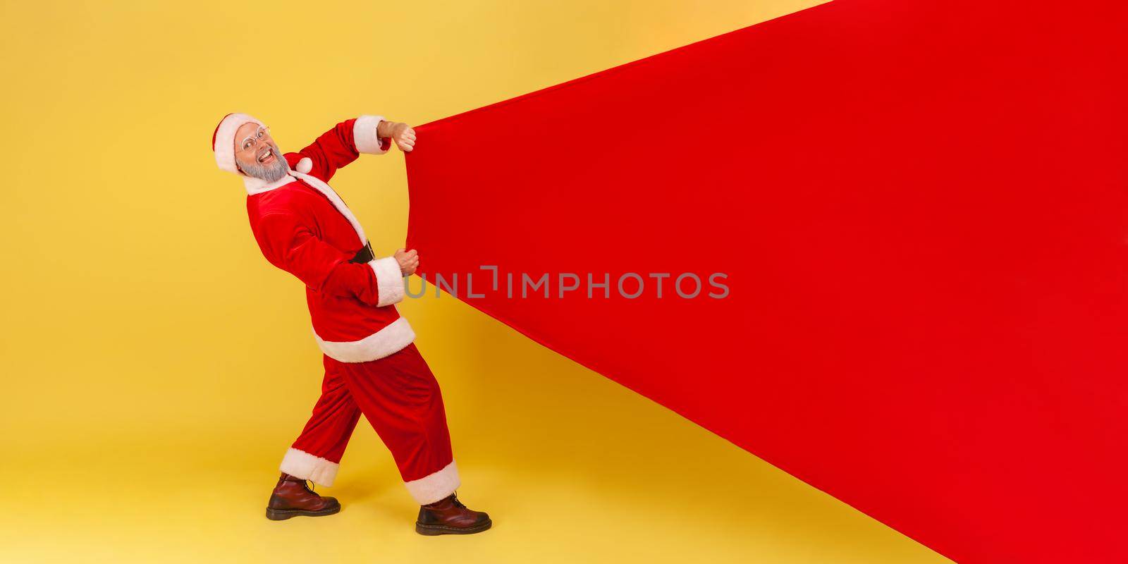 Extremely happy smiling elderly man with gray beard wearing santa claus costume pulling huge bag of gifts, copy space for advertisement. Indoor studio shot isolated on yellow background.