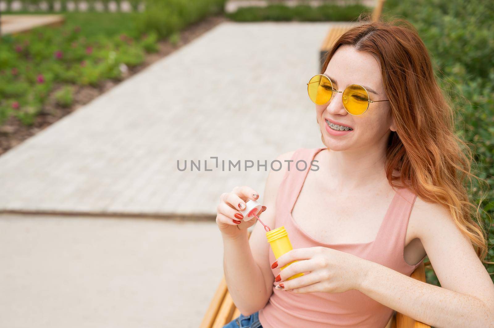Young red-haired woman blowing soap bubbles outdoors. Girl in yellow sunglasses and braces